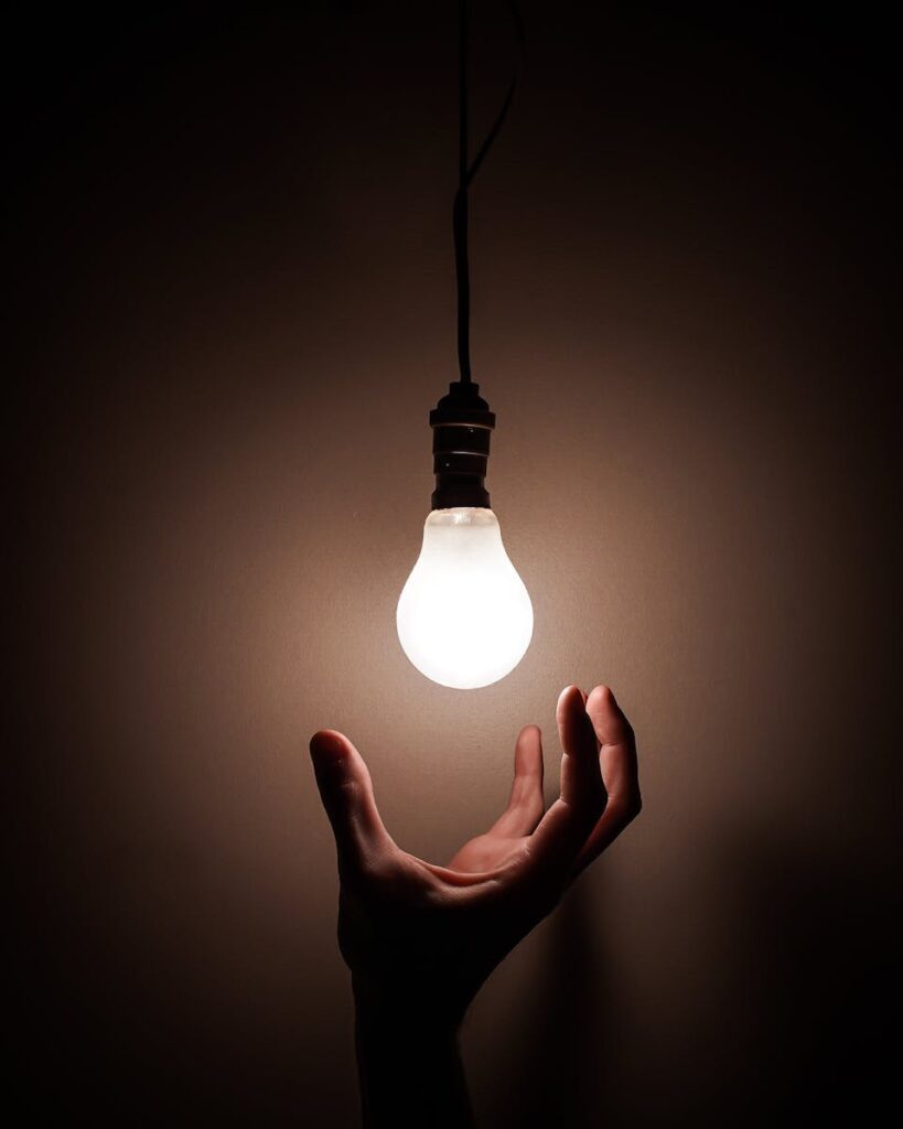 Ruka koja pruža ruku ka užarenoj visećoj sijalici koja simbolizuje kreativnost i poskupljenje električne energije, 