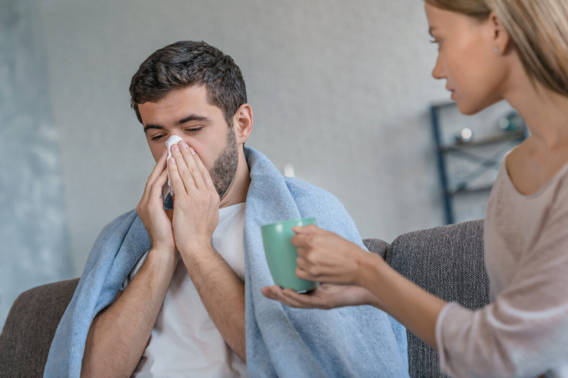Attractive young woman holding cup of warming drink for sick boyfriend while he sneezing